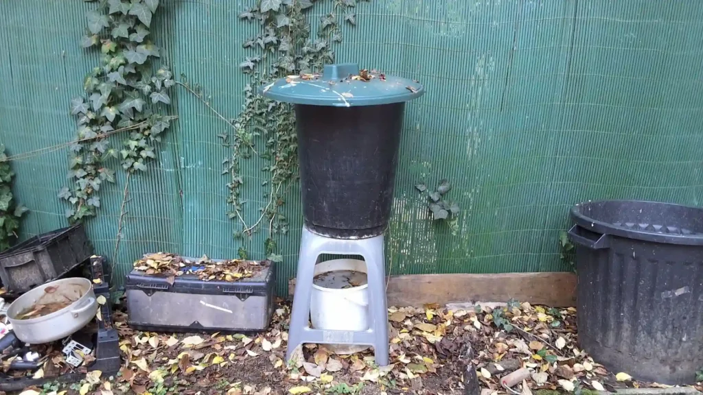 Installation complète d'un lombricomposteur fait maison : un seau noir percé posé sur un tabouret en plastique, avec un seau blanc en dessous pour récupérer l'engrais liquide, le tout placé à l'ombre.
