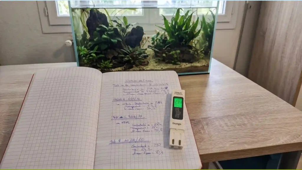 Carnet de mesures d'évaporation aquarium avec conductimètre Dustgo affichant 231 µS, bac naturel low-tech en arrière-plan